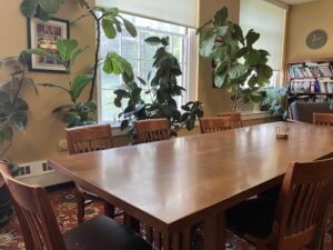 Photo of a long wooden table with chairs. Plants in the background