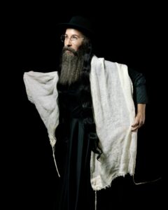 A bearded man stands in front of a black background, looking toward the left.